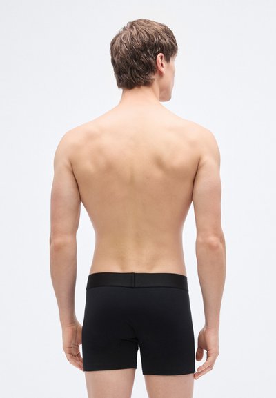 Boxer briefs noirs avec une texture lisse. Ceinture élastique. Coutures visibles et coupe ajustée, montrés de dos sur un fond neutre.