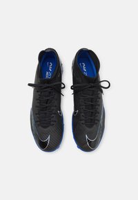 Pár černých fotbalových kopaček s modrými stélkami a podrážkami, se bílými logy Nike swoosh a černými tkaničkami, viděno zeshora na bílém pozadí.
