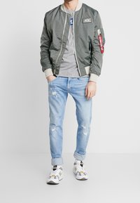 Olivgrön bomberjacka med en krämfärgad ribbad krage, grå t-shirt, slitna ljusblå jeans och mönstrade vita sneakers.