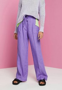 Osoba ubrana w szerokie lawendowe spodnie z zielonymi kieszeniami, lawendowy sweter z dzianiny i czarne klapki na tle różowym.