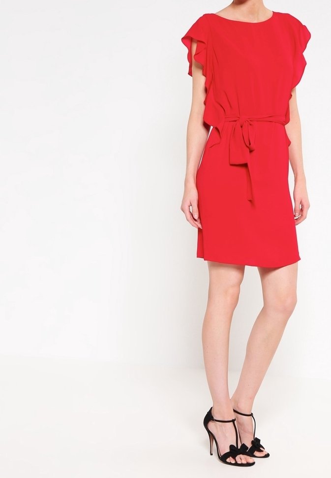 Femme portant une robe rouge mi-longue avec des manches à volants et une ceinture à la taille, associée à des sandales noires à talons hauts ornées de nœuds.