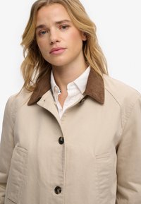 Mujer rubia con chaqueta beige abotonada con cuello marrón sobre camisa blanca, mirando al frente con expresión neutral sobre fondo blanco.