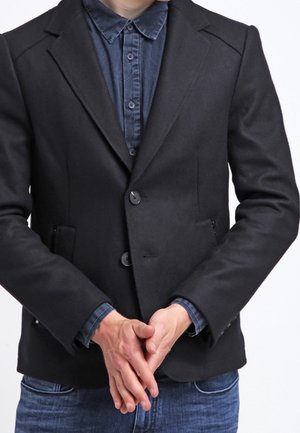 Persoon die een zwart colbert draagt over een blauw overhemd met knopen, met de handen samengevouwen voor zich, gekleed in een blauwe spijkerbroek tegen een witte achtergrond.