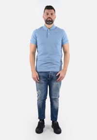 Polo shirt azzurro chiaro con colletto, maniche corte e un logo sottile. Indossato con jeans blu strappati e sneaker nere.