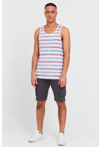 Man standing wearing a striped tank top, dark cargo shorts, white sneakers, and an earring, against a plain white background.