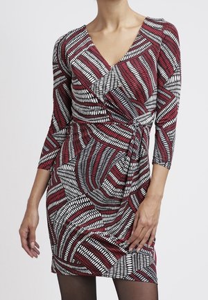 Mujer con un vestido cruzado ajustado de mangas 3/4, con un patrón geométrico en rojo, blanco y negro y un detalle de nudo lateral.