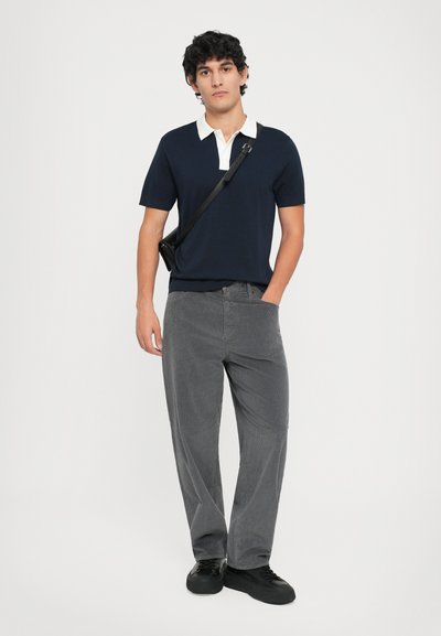 Marineblå polo skjorte med kremfarget krage, kombinert med grå kordfløyelsbukser og sorte joggesko. Modellen holder en svart crossover-veske, stående foran en ensfarget bakgrunn.