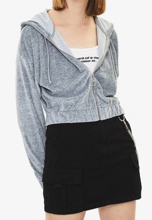 Nainen, jolla on harmaa vetoketjullinen lyhyt huppari valkoisen graafisen topin päällä ja musta minihame, jossa on taskut ja ketjulisäke.