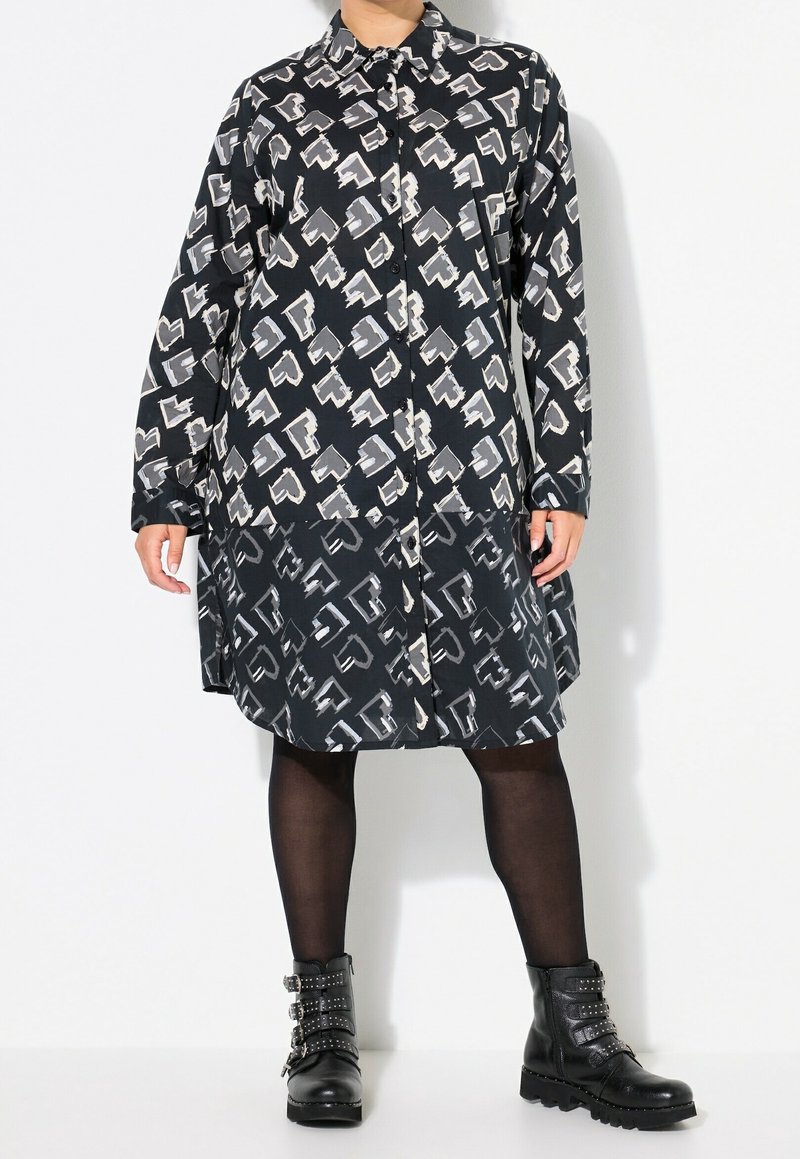 Personne portant une robe à boutons à motif abstrait noir et blanc, des collants noirs et des bottines noires cloutées, debout devant un fond blanc.