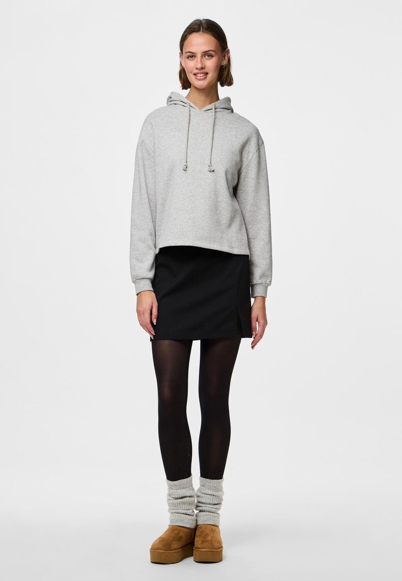 Mladá žena stojí, má na sobě světle šedou mikinu s kapucí, černou mini sukni, černé punčocháče, šedé návleky na nohy a hnědé platformové boty na bílém pozadí.