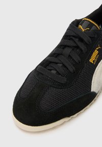 Zapatilla negra con materiales de malla y ante, con cordones negros, suela color crema y un pequeño logo dorado en el lateral.