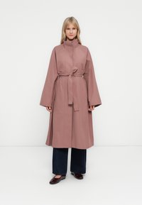 Femme portant un trench-coat mauve ceinturé sur un jean large noir et des ballerines bordeaux, se tenant devant un fond blanc uni.
