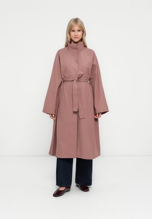 Donna che indossa un trench coat mauve con cintura sopra dei jeans a gamba larga scuri e delle scarpe basse borgogna, in piedi contro uno sfondo bianco semplice.