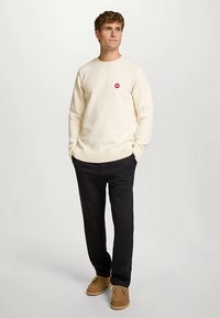 Crèmekleurige gebreide trui met geribbelde manchetten, met een klein rood logo. Gecombineerd met zwarte broek en lichtbruine schoenen.