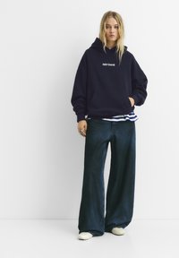 Námornícka oversized mikina s textom „laid-back“, skombinovaná s voľnými tmavými denimovými džínsami a béžovými topánkami, oblečená cez pruhovanú košeľu.