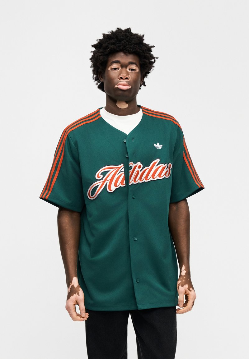 Maillot de sport vert avec manches rayées orange et logo "Adidas" brodé. Présente une fermeture à boutons sur le devant et une coupe décontractée.