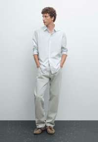 Lichtgrijs button-up shirt gecombineerd met lichtgrijze broek en bruine moccasinschoenen. Model staat met handen in de zakken tegen een neutrale achtergrond.
