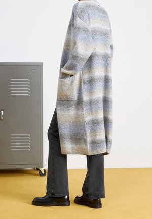 Personne portant un long cardigan rayé et duveteux avec des poches, un jean noir et des chaussures noires, debout à côté d'un casier métallique gris sur un sol jaune.