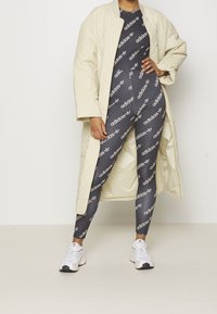 Kremowy pikowany długi płaszcz na czarnych legginsach z białym wzorem logo Adidasa, połączony z białymi butami sportowymi na jednolitym tle.