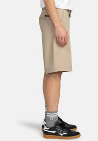 Pantaloni corti chino beige realizzati in cotone, con taglio dritto, tasche laterali e etichetta in vita. Indossati con sneakers nere e calzini grigi.