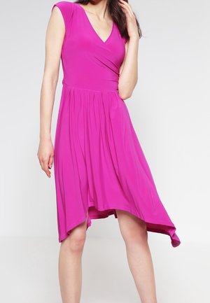 Woman wearing a sleeveless, bright pink asymmetrical hem dress with a V-neckline and gathered waist, standing against a plain background.