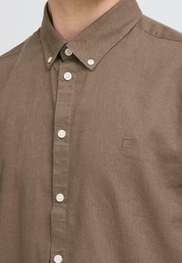 Chemise marron boutonnée avec un tissu texturé, dotée d'un col et de six boutons blancs. Comprend un petit logo brodé sur la poitrine.