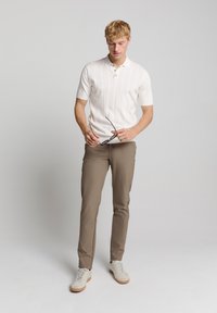 Witte polo met korte mouwen, voorzien van een textuurpatroon en een drieknoopsluiting, gecombineerd met beige slim-fit broek en off-white sneakers.