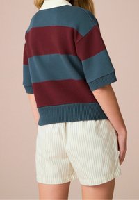 Sweter w paski w kolorach morskim i bordowym, z prążkowanym dekoltem i krótkimi rękawami, zestawiony z białymi szortami w cienkie paski.