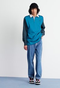 Polo a maniche lunghe color turchese con maniche scure a contrasto, colletto bianco, abbinato a jeans azzurri chiari e sneaker nere con accenti rosa.