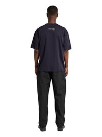 Mężczyzna stoi tyłem, ubrany w ciemnogranatowy T-shirt z małym białym napisem przy szyi i luźne czarne spodnie na jednolitym białym tle.