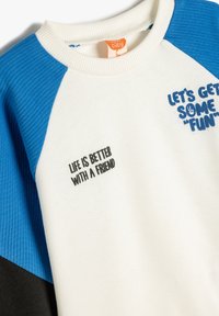 Sweatshirt i hvid, blå og sort. Har ribbet detaljer, legende grafik og tekst: "LAD OS HAVE NOGLE SJOVE TIDER" og "LIVET ER BEDRE MED EN VEN."