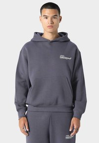 Šedá mikina s kapucí vyrobená z měkké látky, s přední kapsou a vyšitým textem na hrudi. Volný střih s vroubkovanými manžetami.