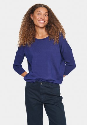 Navyblauwe gebreide trui met een relaxte pasvorm en driekwartmouwen, gecombineerd met donkere broek, met een gladde textuur en casual ontwerp.