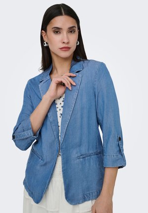 Donna con lunghi capelli scuri indossa un blazer in denim azzurro chiaro sopra una maglia bianca floreale, posando con la mano vicino al colletto su uno sfondo semplice.