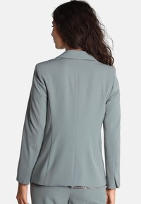 Blazer grigio chiaro realizzato in tessuto liscio. Presenta una vestibilità sartoriale, revers affilati e maniche lunghe. Design semplice senza motivi visibili.