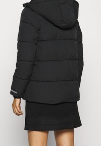 Czarna kurtka puchowa z kapturem, z pikowanymi sekcjami i elastycznymi mankietami. Detale z logo na rękawie oraz teksturowane wykończenie.
