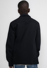 Zwarte, gekraagde jas met knoopmanchetten, gemaakt van een gladde stof; zichtbaar vanaf de achterkant, met een rechte pasvorm en minimalistisch ontwerp.