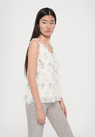 Blusa leggera color crema con motivo floreale, dettagli arricciati e spalline regolabili; abbinata a pantaloni metallici argento con texture.