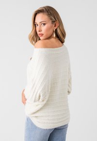 Suéter sin hombros de color blanco roto con un patrón texturizado y acanalado. Presenta mangas sueltas y un corte holgado, combinado con jeans de un azul claro.