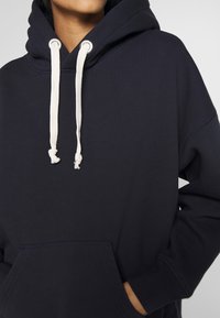 Mörkblå huvtröja i mjukt tyg med en stor framficka, vita dragsnören och en avslappnad passform med ribbade muddar och fåll.