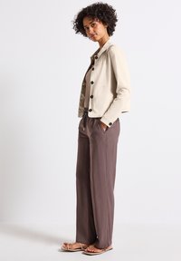 Ljus beige kort jacka med svarta knappar, kombinerad med lösa bruna byxor. Sandaler har tunna remmar och fyrkantiga tår. Lockigt hår.