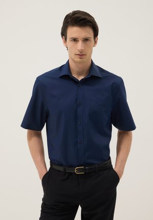 Jonge man met donker haar, die een marineblauw kort mouwen overhemd met knopen draagt en een zwarte broek met een zwarte riem, handen in de zakken.