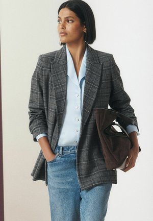 Femme portant un blazer à carreaux gris, une chemise bleu clair boutonnée et un jean, tenant un sac à main marron, regardant sur le côté avec une main dans sa poche.