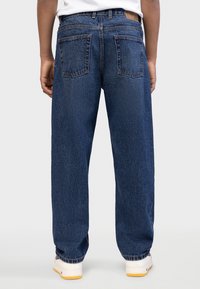 Τζιν παντελόνια, μπλε χρώμα, άνετη εφαρμογή. Διαθέτουν δύο πίσω τσέπες και ραμμένα τελειώματα. Λευκά αθλητικά παπούτσια με κίτρινες λεπτομέρειες ορατές στο κάτω μέρος.