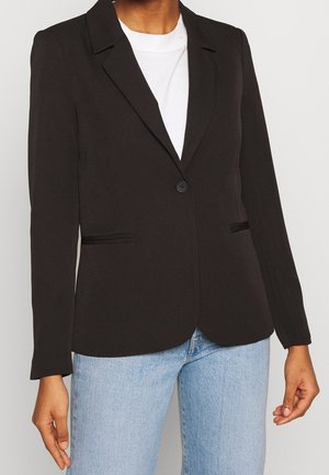 Frau trägt einen enganliegenden schwarzen Blazer über einem weißen Hemd und hellblauen Jeans, steht mit entspannten Armen seitlich am Körper.