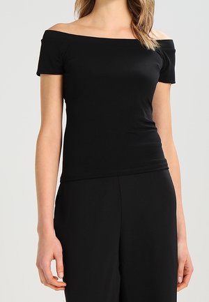 Žena nosící černý přiléhavý top s odhalenými rameny a krátkými rukávy, černé vysoké kalhoty, stojí před jednotným pozadím.