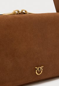 Καφέ τσάντα χειρός από σουέντ με ομαλή υφή, διακοσμημένη με πόρπη από χρυσό μέταλλο και χαρακτηριστικό λογότυπο στο μπροστινό μέρος.