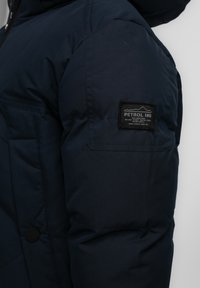 Navyblaue gepolsterte Jacke mit strukturierter Oberfläche, ausgestattet mit einem Logo-Patch am Ärmel und einer Knopfleiste vorne.