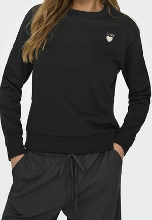 Mujer que lleva una sudadera negra con un pequeño corazón blanco y el texto "LOVE" en el pecho, combinada con pantalones oscuros con cordón.