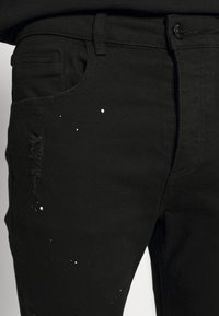 Jeans negros con detalles desgastados y salpicaduras de pintura blanca. El tejido es mezclilla con una textura suave; incluye bolsillos en la parte delantera.